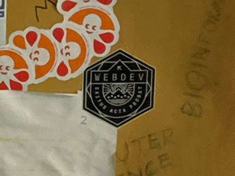 A black hexagonal sticker that reads WEBDEV: EXITUS ACTA PROBAT, affixed to the corner of a sheet of paper against a light brown background