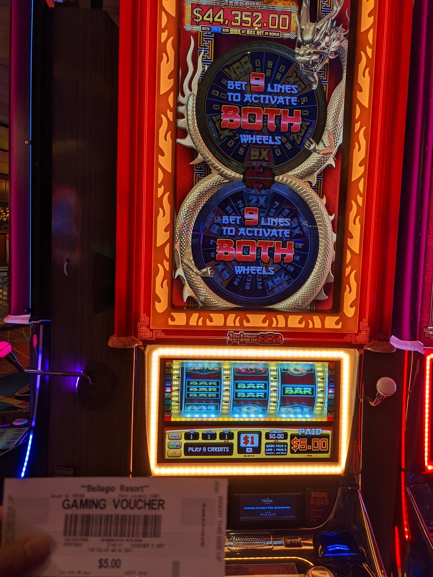 A $5.00 'gaming voucher' held in front of a slot machine with a golden dragon circling two wheels, which is indicating that it costs $1 to pay and just paid out $5.00